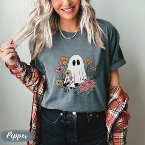 K&ouml;nnte beinhalten: Ein dunkelgraues T-Shirt mit einem wei&szlig;en Geist, der von bunten Blumen umgeben ist. Der Geist hat zwei schwarze Augen. Der Text "Pepper" ist in der linken unteren Ecke des Bildes sichtbar.