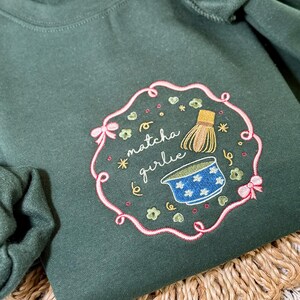 Puede incluir: Sudadera verde oscuro con un dise&ntilde;o bordado. El dise&ntilde;o presenta el texto "matcha girlie" en cursiva, un cuenco de matcha y un batidor, rodeados por un borde rosa con lazos y detalles florales. La sudadera se muestra sobre una superficie tejida.