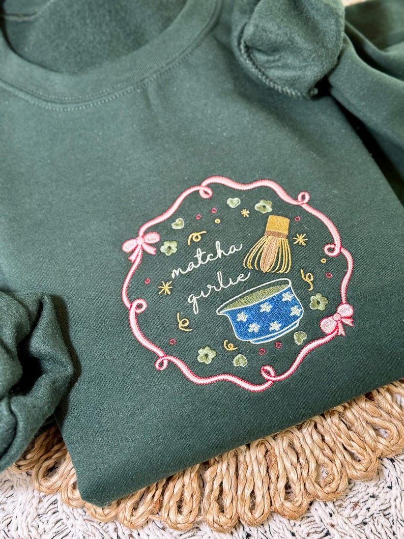 Puede incluir: Sudadera verde oscuro con un dise&ntilde;o bordado. El dise&ntilde;o presenta el texto "matcha girlie" en cursiva, un cuenco de matcha y un batidor, rodeados por un borde rosa con lazos y detalles florales. La sudadera se muestra sobre una superficie tejida.