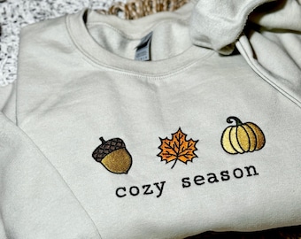 May include: A light gray sweatshirt with an embroidered design of an acorn, a leaf, and a pumpkin. The text "cozy season" is embroidered below the design.