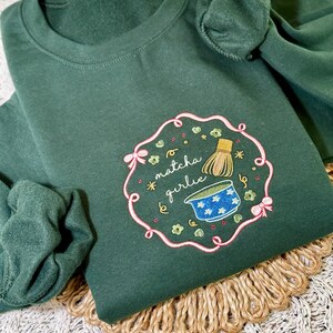 Puede incluir: Sudadera verde oscuro con un dise&ntilde;o bordado. El dise&ntilde;o presenta el texto "matcha girlie" en cursiva, junto con un cuenco de matcha, un batidor y detalles florales. El dise&ntilde;o est&aacute; enmarcado por un borde rosa con lazos.