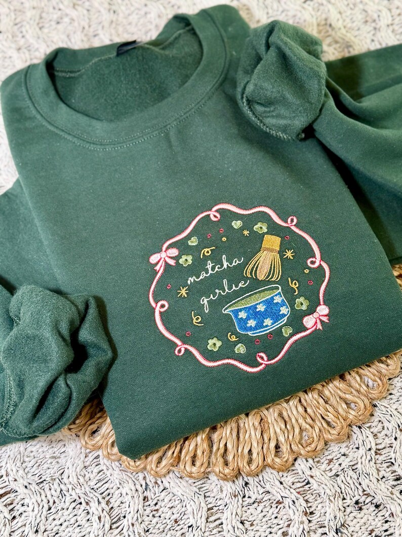 Puede incluir: Sudadera verde oscuro con un dise&ntilde;o bordado. El dise&ntilde;o presenta el texto "matcha girlie" en cursiva, junto con un cuenco de matcha, un batidor y detalles florales. El dise&ntilde;o est&aacute; enmarcado por un borde rosa con lazos.