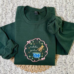 Puede incluir: Sudadera verde oscuro con cuello redondo y mangas largas. La parte delantera presenta un dise&ntilde;o bordado con las palabras "matcha girlie", un cuenco de matcha y un batidor. El dise&ntilde;o est&aacute; enmarcado por un borde floral rosa.
