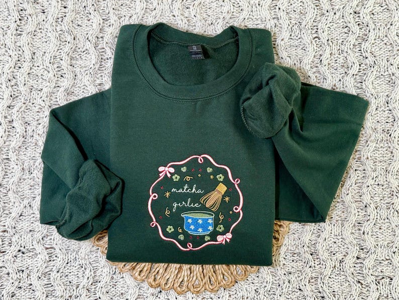 Puede incluir: Sudadera verde oscuro con cuello redondo y mangas largas. La parte delantera presenta un dise&ntilde;o bordado con las palabras "matcha girlie", un cuenco de matcha y un batidor. El dise&ntilde;o est&aacute; enmarcado por un borde floral rosa.