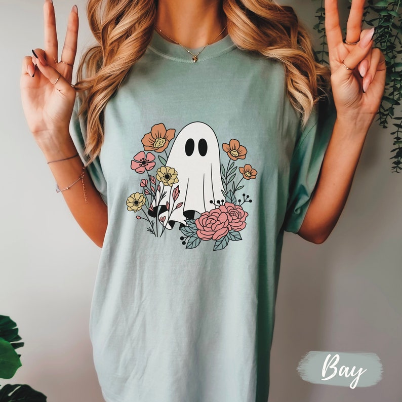 K&ouml;nnte beinhalten: Ein hellblaues T-Shirt mit einem wei&szlig;en Geist und einem floralen Design. Der Geist hat zwei schwarze Augen und ist von rosa, gelben und orangefarbenen Blumen umgeben.