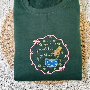 Puede incluir: Una sudadera verde oscuro con un dise&ntilde;o bordado. El dise&ntilde;o presenta las palabras "matcha girlie" en cursiva, un cuenco azul, un batidor de bamb&uacute; y elementos decorativos dentro de un marco rosa y verde.