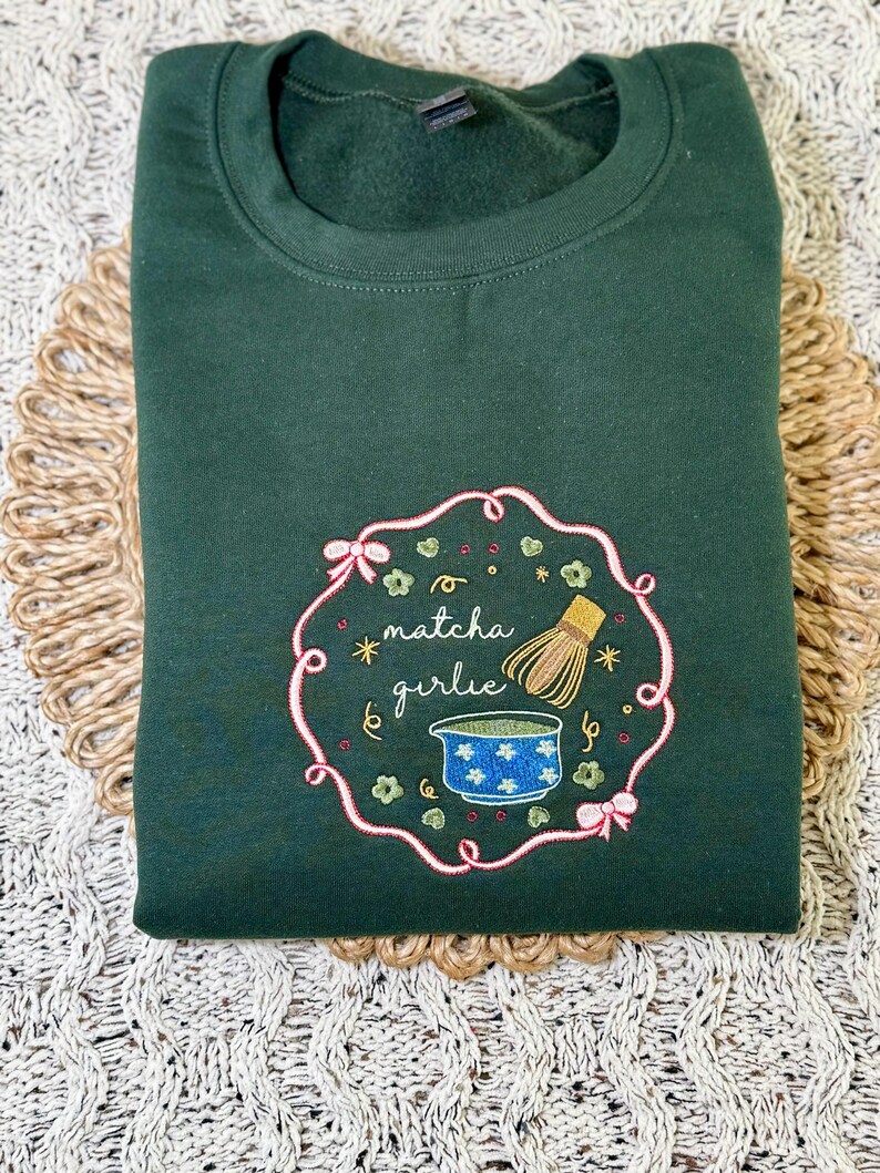 Puede incluir: Una sudadera verde oscuro con un dise&ntilde;o bordado. El dise&ntilde;o presenta las palabras "matcha girlie" en cursiva, un cuenco azul, un batidor de bamb&uacute; y elementos decorativos dentro de un marco rosa y verde.