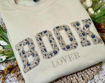 Puede incluir: Una sudadera de cuello redondo en color beige con la palabra BOOK en letras grandes de estilo bloque rellenas con un patrón floral azul oscuro sobre fondo claro. Debajo de la palabra BOOK, la palabra LOVER está bordada en una fuente beige fina.