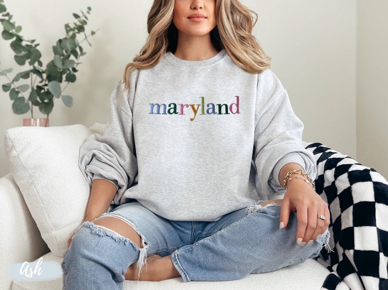 May include: A gray sweatshirt with the word "maryland" printed on the front in colorful letters. The sweatshirt is being worn by a person sitting on a white couch.