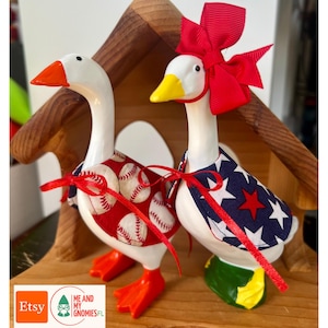 Pode incluir: Duas figuras decorativas de gansos. Um ganso usa uma capa vermelha com tema de beisebol, o outro uma capa azul com estrelas brancas e uma estrela vermelha. Ambos têm corpos brancos, pés e bicos laranja e laços vermelhos. Eles são exibidos em uma estrutura de madeira.