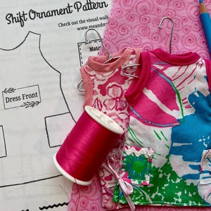 May include: A DIY craft kit for making a pink shift dress ornament. The kit includes a pattern, fabric scraps, thread, and a hanger. The pattern includes instructions for making the dress and a dress front template.