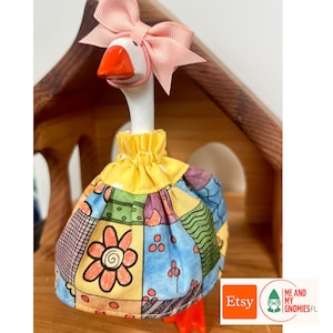 May include: A white goose figurine with an orange beak, wearing a light pink ribbon bow on its head and a gathered yellow-collared dress featuring a multi-colored patchwork pattern of flowers, dots, and wavy lines in blue, orange, and green.