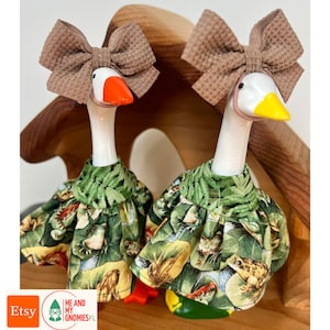 May include: Two decorative goose figurines with white bodies, orange beaks, and yellow feet. They wear green and yellow frog-themed dresses and brown bow headbands. The Etsy logo is visible in the bottom left corner.