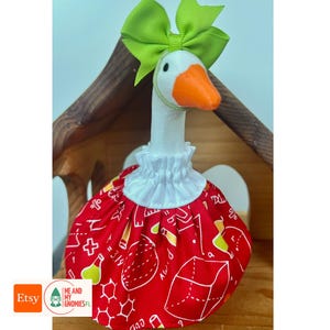 May include: A white goose figurine wearing a bright lime green ribbon bow on its head and a red fabric dress featuring a white print of mathematical symbols, geometric shapes, and science beakers, finished with a white ruffled collar.