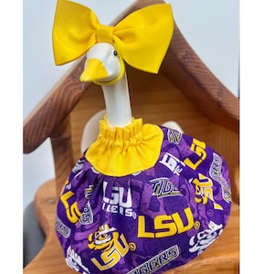 May include: A white ceramic goose figurine wearing a bright yellow ribbon bow on its head and a gathered yellow collar. The goose is dressed in a purple fabric skirt featuring a repeating white and yellow LSU Tigers logo and tiger head print.