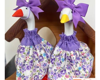 May include: Two white goose figurines wearing matching purple fabric dresses with a floral pattern of purple, blue, and pink flowers. Each goose has a purple ribbon bow on its head, a ruffled purple collar, and a lace hem. One has an orange beak, the other yellow.