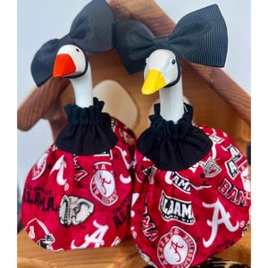 May include: Two white goose figurines wearing black grosgrain ribbon bows on their heads and red fabric outfits featuring the University of Alabama Crimson Tide logo, elephant mascot, and text. One goose has an orange beak and the other a yellow beak.