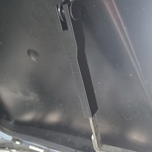 May include: Close-up of a black metal hinge mechanism with a roller. The hinge is attached to a black surface and is in an open position.