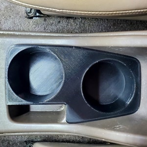 May include: A close-up of a black plastic cup holder with two circular openings. The cup holder is installed in a car's center console.