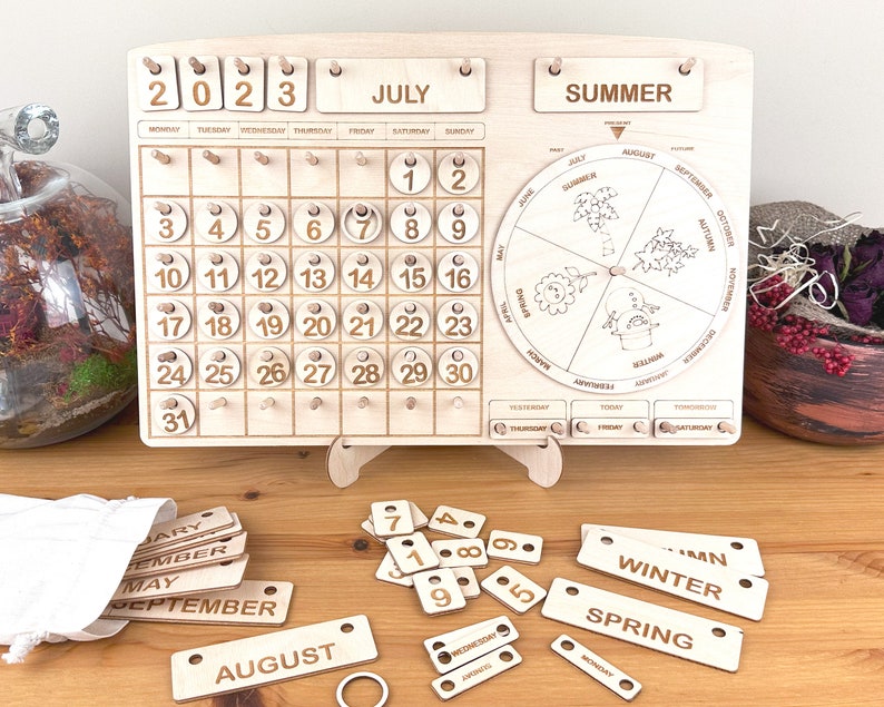 May include: Wooden calendar with a monthly calendar, a seasonal wheel, and separate pieces for days, months, and seasons. The calendar is set to July 2023. The seasonal wheel shows the seasons of the year with illustrations of flowers, leaves, and snow.