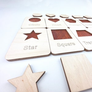 May include: Wooden puzzle pieces with cutouts of different shapes, including a circle, heart, hexagon, star, square, and triangle. Each piece has the name of the shape written below the cutout.