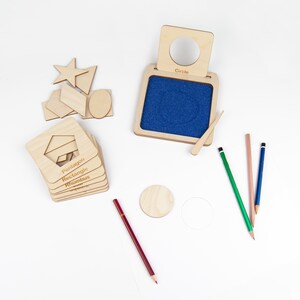 May include: Wooden geometric shapes learning toy with a blue felt board and a wooden stylus. The shapes include a circle, star, pentagon, rectangle, rhombus, and a few other shapes. The shapes are labeled with their names. There are also four colored pencils on the white surface.