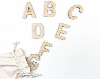 May include: Wooden letters A through G are scattered on a white surface. A small white drawstring bag is in the lower left corner of the image.