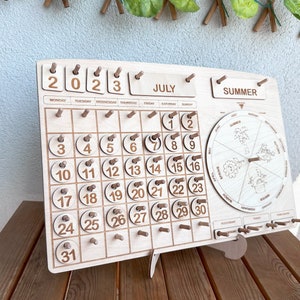 May include: Wooden calendar with a July 2023 calendar, a summer season wheel, and a daily tracker. The calendar has a light wood finish and the numbers are in brown.