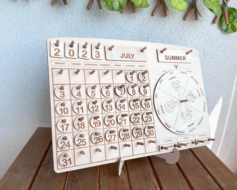 May include: Wooden calendar with a July 2023 calendar, a summer season wheel, and a daily tracker. The calendar has a light wood finish and the numbers are in brown.