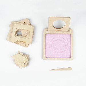 May include: A wooden tray with a pink sand surface and a cutout circle. The tray has a wooden handle with the word "Circle" printed on it. There are also wooden shapes, including a rectangle and a diamond, in the image.