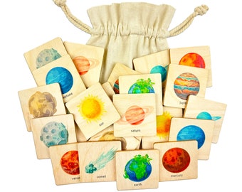 Op de afbeelding: Een set houten planeten- en ruimtethema-tegels in een trekkoordtas. De tegels bevatten illustraties van planeten, de zon, een komeet en de woorden "zon", "aarde", "mercurius", "venus", "mars", "jupiter", "saturnus", "uranus", "neptunus" en "komeet".