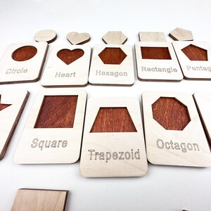 May include: Wooden learning cards with different shapes cut out. The shapes include a circle, heart, hexagon, rectangle, pentagon, square, trapezoid, and octagon. Each card has the name of the shape printed on it.
