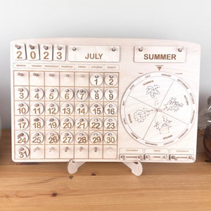May include: Wooden calendar with a monthly calendar grid for July 2023, a rotating wheel with the seasons of the year, and a day of the week indicator. The calendar is made of light wood and has a natural finish.