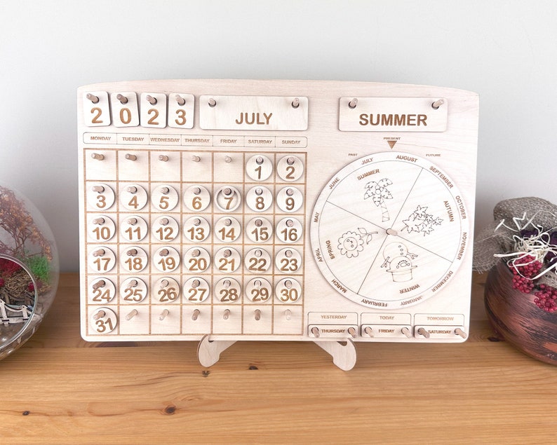 May include: Wooden calendar with a monthly calendar grid for July 2023, a rotating wheel with the seasons of the year, and a day of the week indicator. The calendar is made of light wood and has a natural finish.