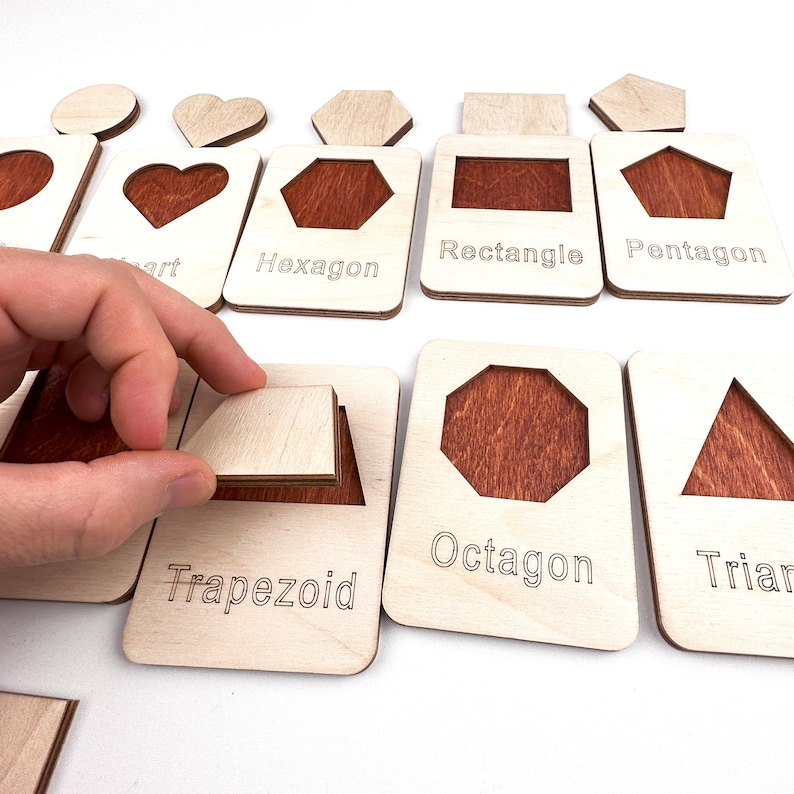 May include: Wooden educational toy with different shapes, including a heart, hexagon, rectangle, pentagon, trapezoid, octagon, and triangle. Each shape has a corresponding cutout on a separate piece of wood.