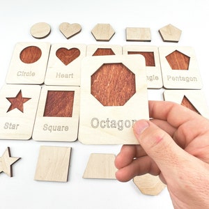 May include: Wooden learning toy with different shapes, including a circle, heart, square, star, octagon, pentagon, and triangle. Each shape has a cutout and a corresponding wooden piece to fit inside.