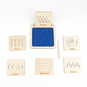 May include: Wooden tracing board with a blue felt surface and six different wooden tiles with various line patterns: diagonal, vertical, spiral, curved, horizontal, and zig-zag. Each tile has a label with the corresponding line pattern name.