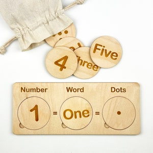 May include: Wooden learning toy with three sections labeled "Number", "Word", and "Dots". The sections have cutouts for number, word, and dot cards. The cards show the number 1, the word "One", and one dot.