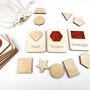 May include: Wooden shape puzzle with a variety of shapes including a heart, hexagon, rectangle, star, circle, and square. The shapes are labeled with their names. The puzzle is made of natural wood and has a light brown color.