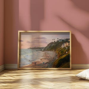 May include: A framed print of a coastal scene. The image features a beach with people, a cliffside with buildings, and the ocean under a sunset sky. The frame is gold-colored, and the print is displayed on a pink wall.