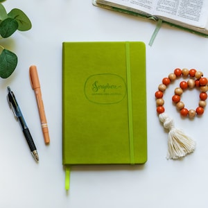 May include: A green journal with the text "Scripture Mapping Mini Journal" on the front cover. The journal is on a white surface with a pen, highlighter, and a string of beads with a tassel.