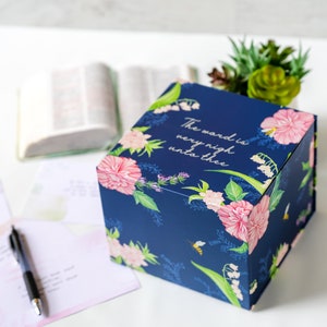 May include: A navy blue gift box with floral and bee illustrations. The box features pink peonies, white flowers, and the text "The word is very nigh unto thee." A pen, open book, and plant are in the background.