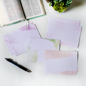 May include: A stack of blank stationery cards with watercolor floral designs in shades of pink, green, and peach. The cards are on a white surface with a pen and a closed book.
