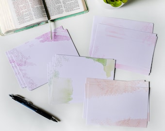 May include: A stack of blank stationery cards with watercolor floral designs in shades of pink, green, and peach. The cards are on a white surface with a pen and a closed book.
