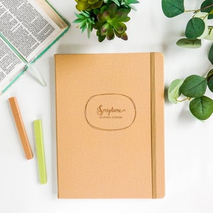 May include: A brown leather-bound journal with a gold inscription that reads "Scripture Mapping Journal". The journal is on a white surface with green plants in the background.