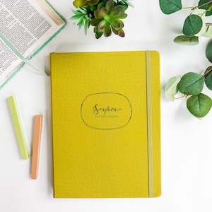 May include: A yellow journal with a green elastic closure. The front cover has a black oval with the text "Scripture Mapping Journal" in a handwritten font.