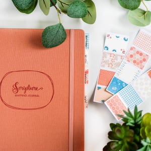 Peut inclure: Un journal de cartographie des Écritures de couleur corail avec les mots "Scripture Mapping Journal" dans une police décorative. Le journal a un élastique assorti et des notes autocollantes à motifs coordonnés.