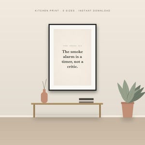 Puede incluir: Una lámina enmarcada con el texto negro 'The smoke alarm is a timer, not a critic.' sobre un fondo beige claro. La lámina se muestra sobre un banco de madera con un pequeño jarrón y una planta en maceta cerca, creando una composición sencilla.