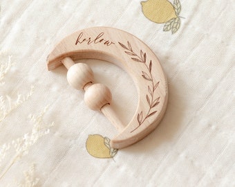 May include: A wooden crescent moon rattle with a leaf design and the name "Harlow" engraved on it. The rattle has wooden beads that slide along a wooden dowel.