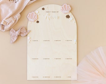 May include: A wooden growth chart with a light brown background and a white arch. The arch has the text "FIRST YEAR OF Aurora" and is decorated with pink and white seashells. The chart has 12 spaces for marking a child's growth, labeled with months and "1 YEAR".